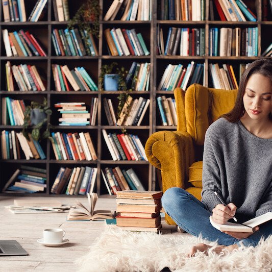Young female Word Boutique style writer in library indoors working sitting on the carpet taking notes in organizer for book thoughtful concentrated